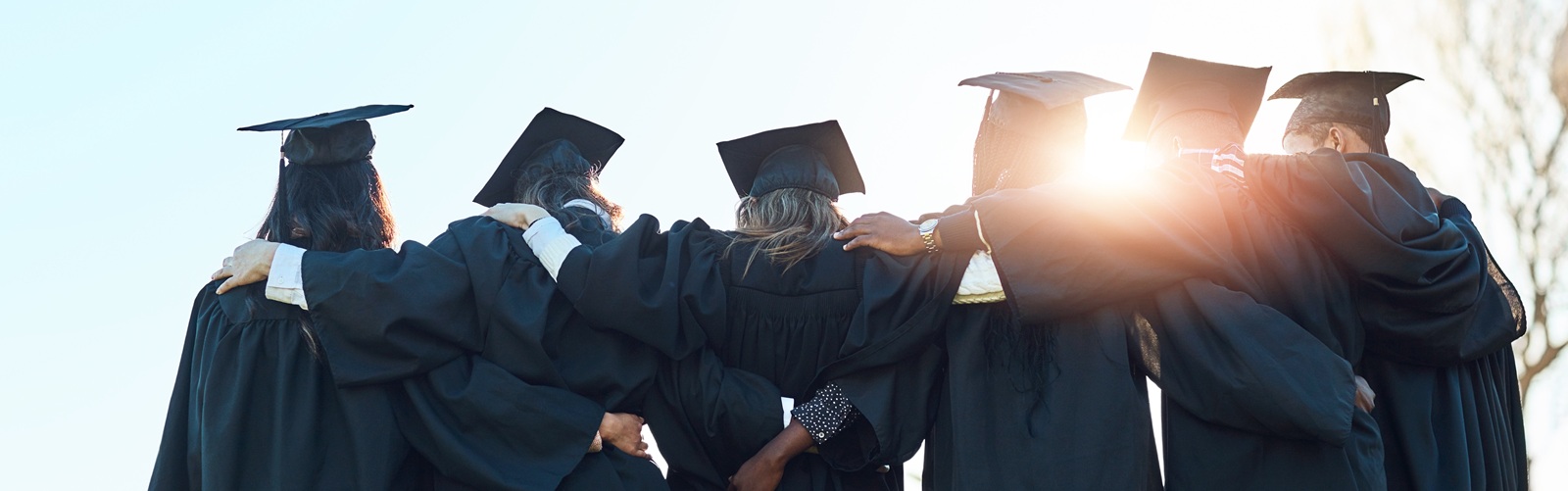group of 6 recent graduates in their cap and gowns standing side by side with arms around each other